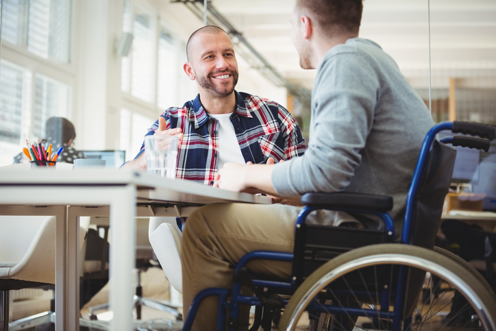 young man in wheelchair works with another young man at an office desk