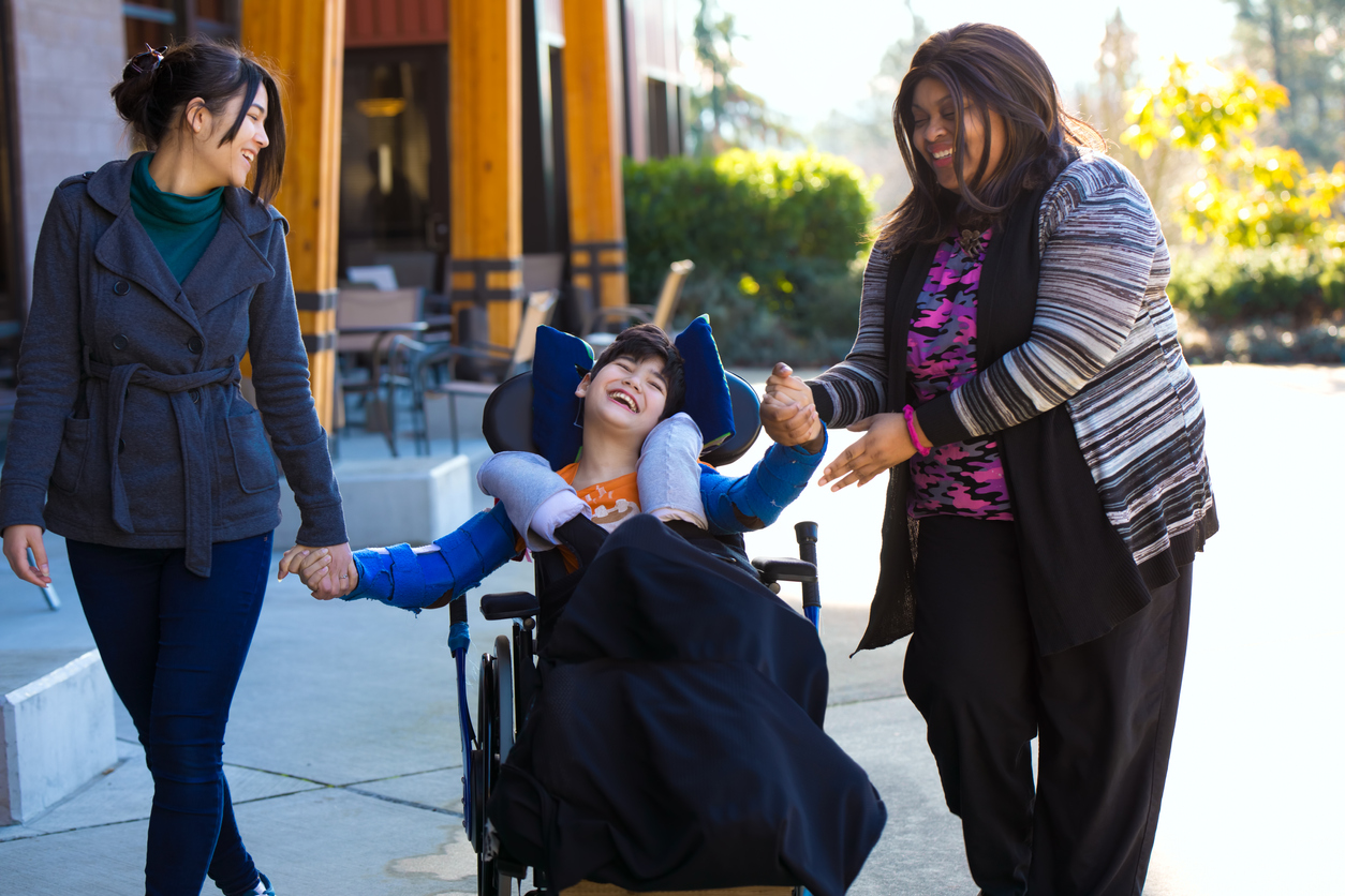 two women help a young boy in wheelchair stroll through an outdoor mall